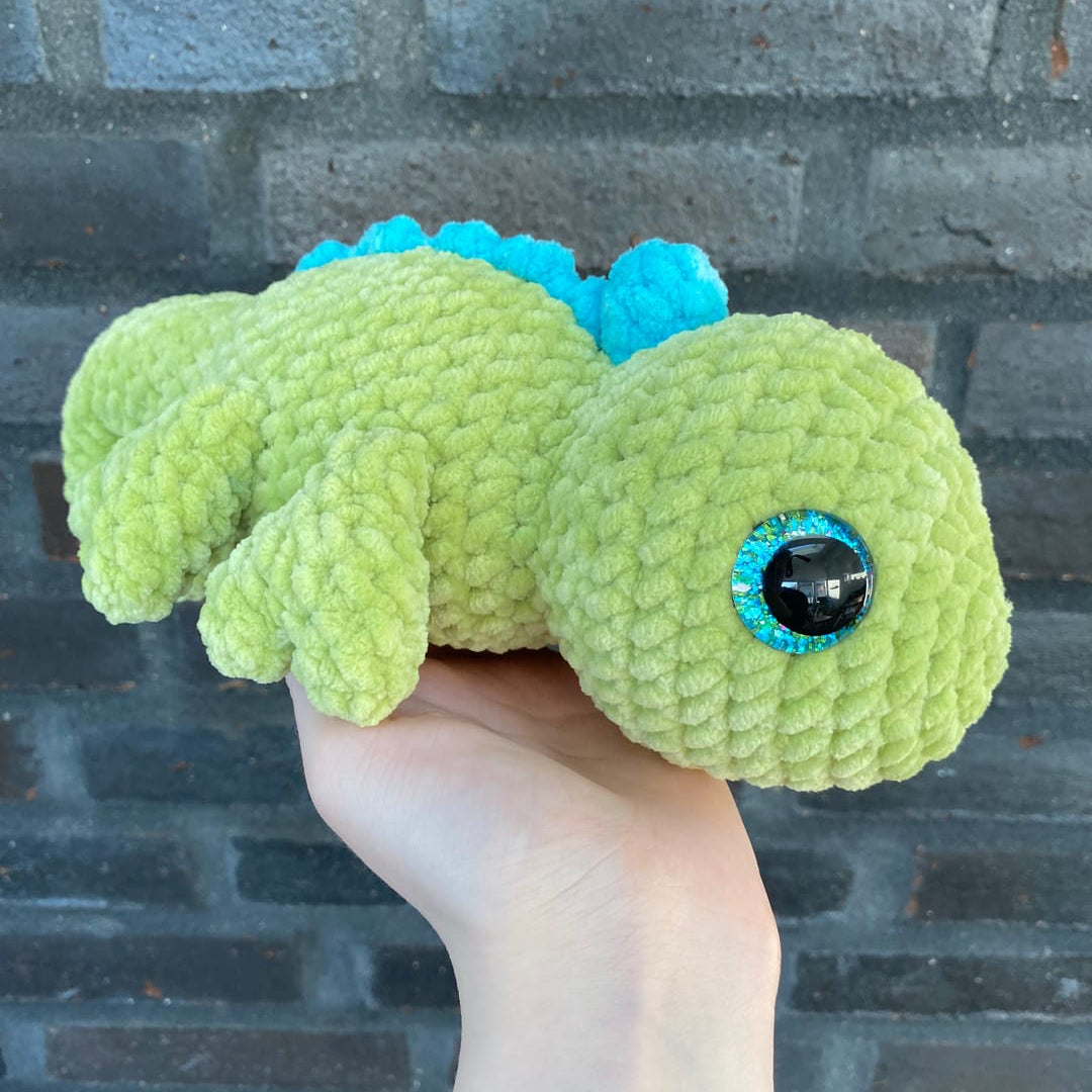 Hand holding a green and blue crochet dragon toy against a brick wall.